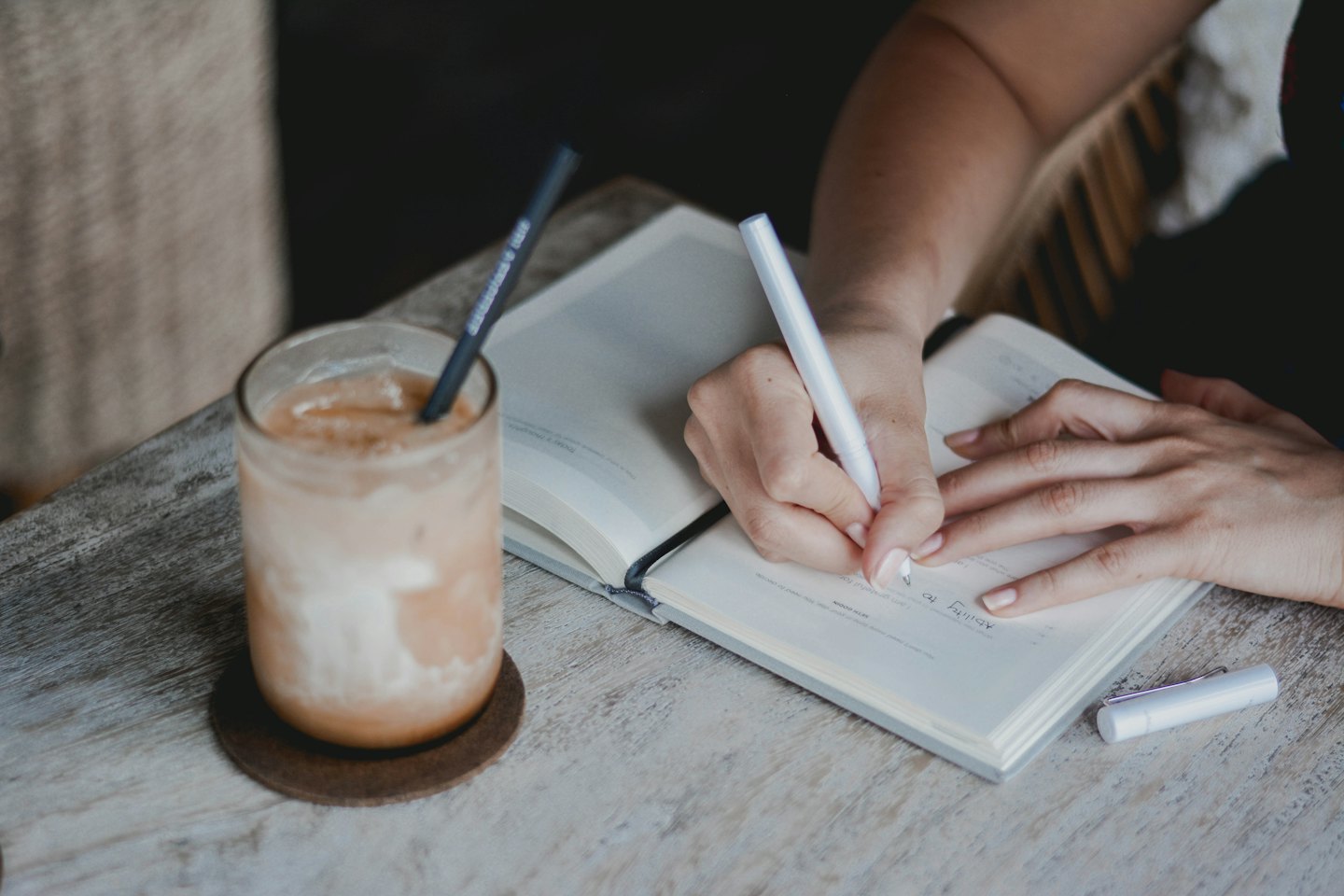 Persona escribiendo en un cuaderno con bolígrafo y café helado.